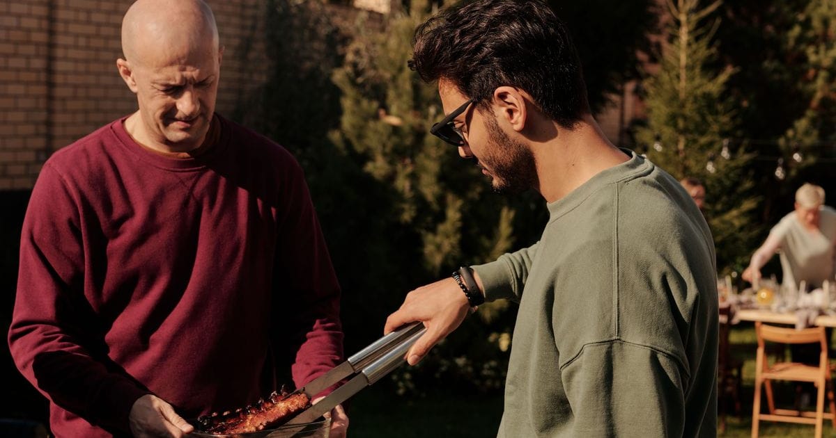 Two dads at a backyard grill, one lifting ribs off with tongs, a family table behind them