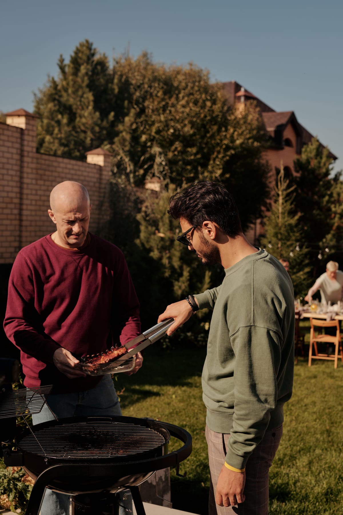 Two dads at a backyard grill in late afternoon light, a family table behind them